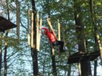 Hochseilgarten Eine Person in einem roten Anzug klettert in einem Hochseilgarten zwischen Bäumen in luftiger Höhe.