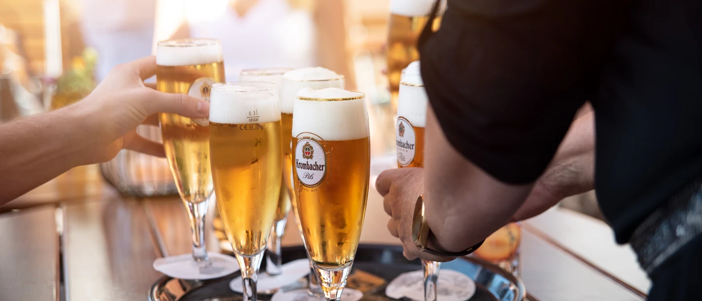 Sechs Personen stoßen mit vollen Biergläsern auf einem Tablett im Freien an.