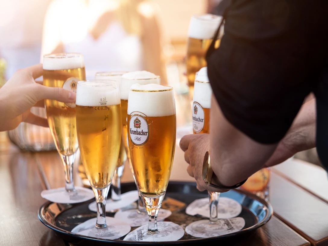 Gaststaette Zum Hoelzchen Tablett mit Bierglaesern.jpg Sechs Personen stoßen mit vollen Biergläsern auf einem Tablett im Freien an.