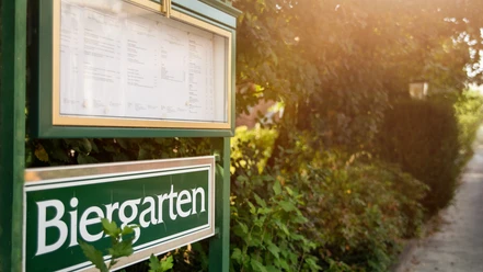 Grünes Schild mit der Aufschrift "Biergarten" an einem sonnigen Pfad, umgeben von üppigem Grün.