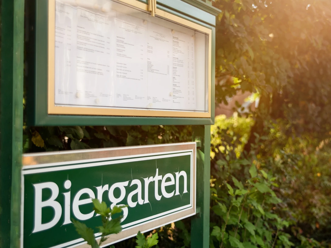Grünes Schild mit der Aufschrift "Biergarten" an einem sonnigen Pfad, umgeben von üppigem Grün.