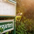 Grünes Schild mit der Aufschrift "Biergarten" an einem sonnigen Pfad, umgeben von üppigem Grün.