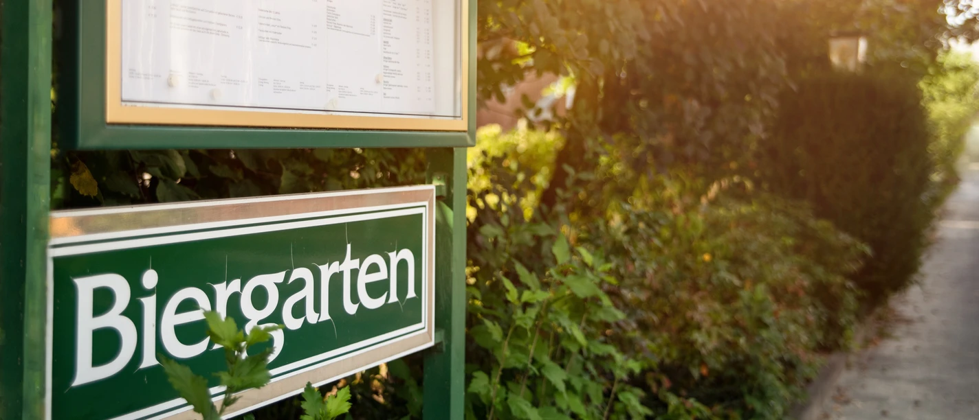 Gaststaette Zum Hoelzchen Biergarten.jpg Grünes Schild mit der Aufschrift "Biergarten" an einem sonnigen Pfad, umgeben von üppigem Grün.