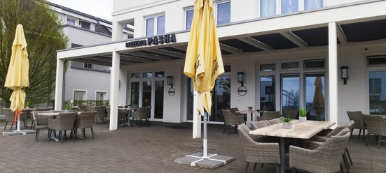 Außenbereich der Pizzeria Pasha mit überdachter Terrasse, Rattanmöbeln und gelben Sonnenschirmen.