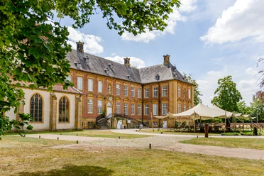 Kloster Marienfeld | Teutoburger Wald | Harsewinkel Barockes Schloss mit Backsteinfassade, großen Fenstern, Zeltpavillon und Garten, umgeben von Bäumen.