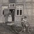 Ein Junge mit Fahrrad steht vor einem traditionellen Fachwerkhaus; ein Paar schaut aus der Tür.