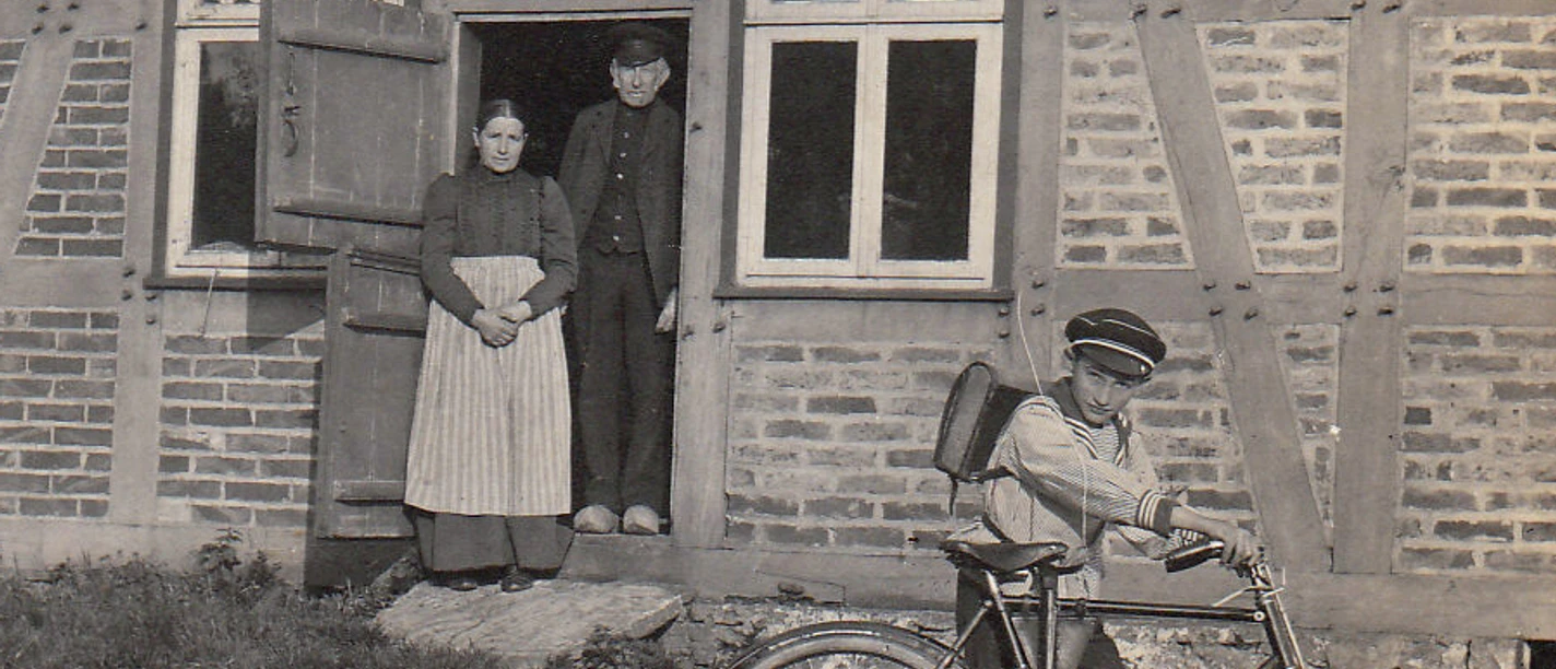 Eggeberg Willi Wesselman mit Schülermütze und Fahrrad um 1912 bearbeitet - Leihgabe von Martin Surmann.jpg Ein Junge mit Fahrrad steht vor einem traditionellen Fachwerkhaus; ein Paar schaut aus der Tür.