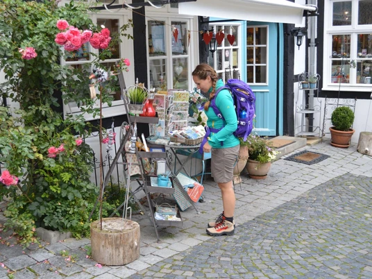 Eine Wanderin mit lila Rucksack betrachtet einen Flohmarktstand in einer gepflasterten Altstadtgasse.