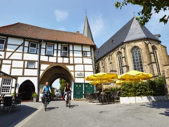 Lengerich_Römer-1_Foto Stadt Lengerich.jpg Fachwerkhaus mit Torbogen und eine Kirche, an deren Platz Fahrräder vorbeifahren.