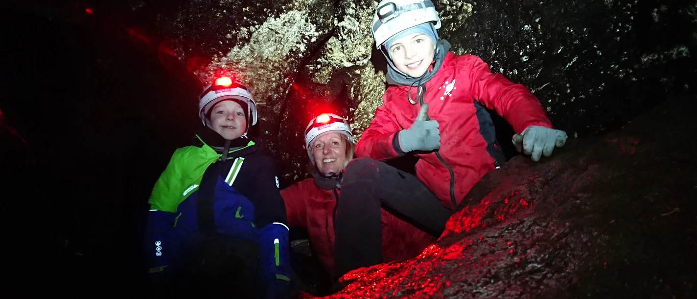 Drie mensen met helmen en hoofdlampen verkennen een donkere grot omgeven door rotswanden.