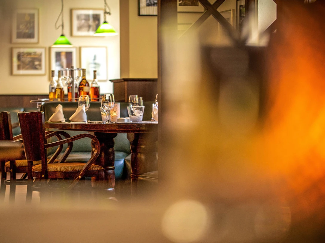 Restaurace Tilia v hotelu Lindenhof Gemütlich gedeckter Tisch in einem Restaurant mit Holzmöbeln, Gläsern, Flaschen und grünen Hängelampen im Hintergrund, warme Atmosphäre.Comfortably laid table in a restaurant with wooden furniture, glasses, bottles and green hanging lamps in the background, warm atmosphere.Útulné prostírání v restauraci s dřevěným nábytkem, sklenicemi, lahvemi a zelenými závěsnými lampami v pozadí, příjemná atmosféra.Przytulne nakrycie stołu w restauracji z drewnianymi meblami, szklankami, butelkami i zielonymi wiszącymi lampami w tle, ciepła atmosfera.Gezellige tafelschikking in een restaurant met houten meubels, glazen, flessen en groene hanglampen op de achtergrond, warme sfeer.Tavola accogliente in un ristorante con mobili in legno, bicchieri, bottiglie e lampade verdi sullo sfondo, atmosfera calda.