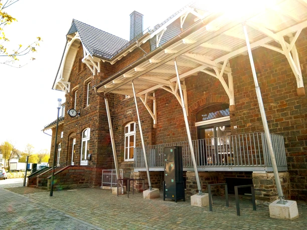 Kultur-Bahnhof-Morsbach Alter Backsteinbahnhof mit Uhr an der Fassade, Holzdach und gepflastertem Vorplatz in Sonnenlicht.