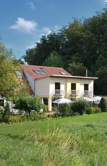 Landgasthof Mücke Marsberg _ 3.jpg Ein zweistöckiges Gebäude mit Terrasse und Sonnenschirmen in grüner, von Bäumen umgebener Landschaft.