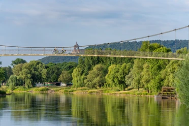 Schiffmühle Minden Zwei Radfahrer überqueren eine Hängebrücke über einem Fluss umgeben von üppiger Vegetation.