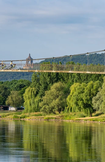 Schiffmühle Minden Zwei Radfahrer überqueren eine Hängebrücke über einem Fluss umgeben von üppiger Vegetation.