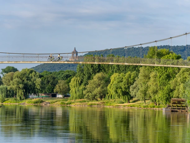 Schiffmühle Minden Zwei Radfahrer überqueren eine Hängebrücke über einem Fluss umgeben von üppiger Vegetation.