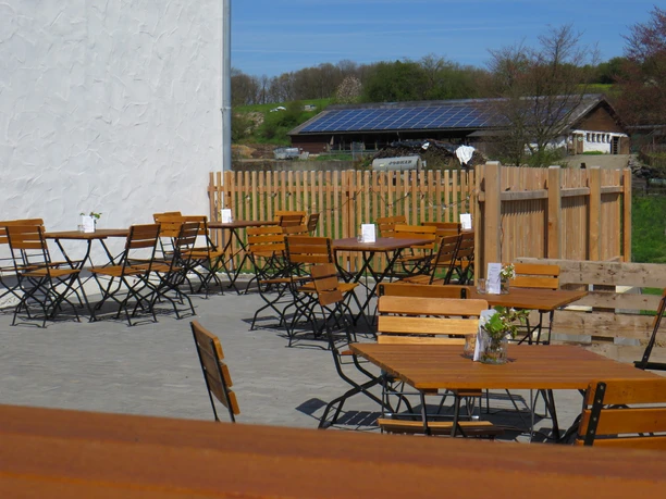 Hofladen Außenbereich Sonniger Außenbereich eines Cafés mit Holztischen und Stühlen, Zaun und ländlicher Landschaft im Hintergrund.