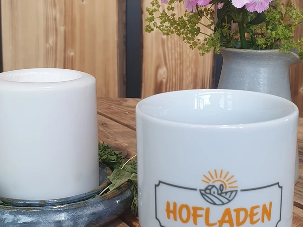 Weiße Tasse mit "Hofladen Ülfetal"-Logo, daneben Kerze und pinke Blumen auf Holztisch.
