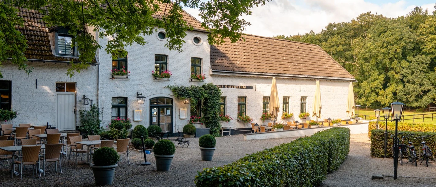Weiß gestrichenes Gebäude im traditionellen Landhausstil, umgeben von grüner Natur. Vor dem Gebäudeeingang stehen zahlreiche Tischgruppen.