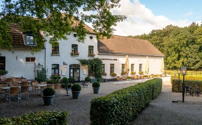 Gut Entenpfuhl Weiß gestrichenes Gebäude im traditionellen Landhausstil, umgeben von grüner Natur. Vor dem Gebäudeeingang stehen zahlreiche Tischgruppen.
