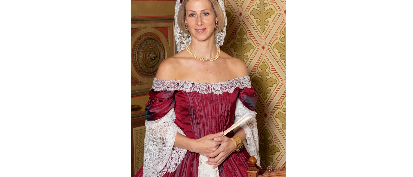 Zeitreise Gräfin Frieda Die bestehende Beschreibung lautet: "Das Bild zeigt die Stadtführerin." Hier ist die optimierte Version: "Stadtführerin in historischer Tracht vor kunstvollem, antikem Hintergrund stehend."