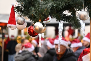 Merry X - Mas everybody! Weihnachtlich geschmückter Tannenbaum, Menschen mit Weihnachtsmützen im Hintergrund.