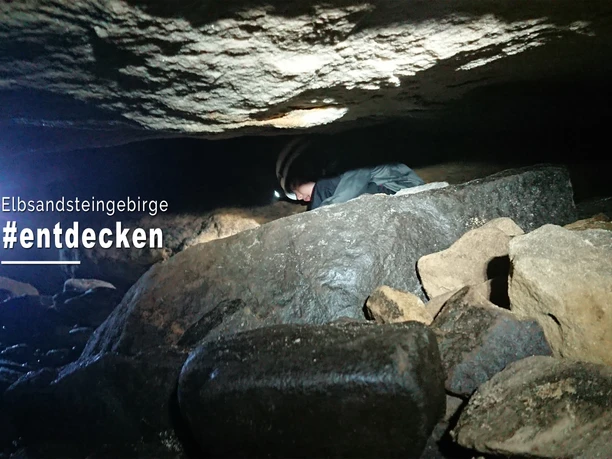 Person crawls with a headlamp through a dark cave in the Elbe Sandstone Mountains, surrounded by rocks and stones.