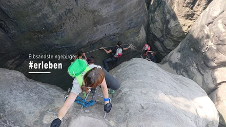 Klettersteigtouren Elbsandsteinguides Personen klettern in einer steilen Felswand im Elbsandsteingebirge, tragen Rucksäcke und nutzen Seile; die Atmosphäre wirkt abenteuerlich.