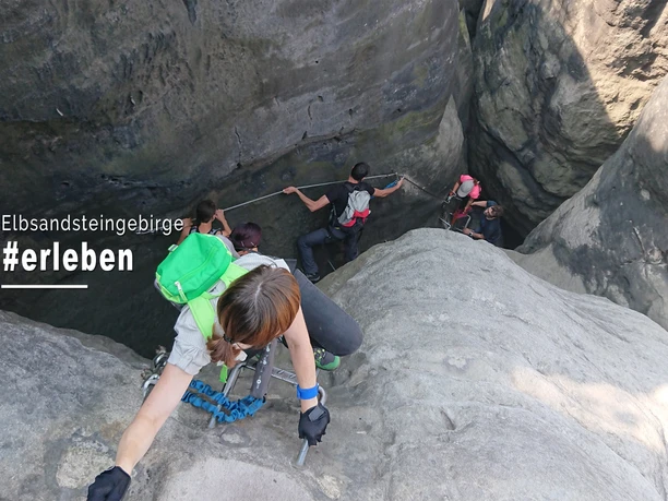 Klettersteigtouren Elbsandsteinguides Personen klettern in einer steilen Felswand im Elbsandsteingebirge, tragen Rucksäcke und nutzen Seile; die Atmosphäre wirkt abenteuerlich.