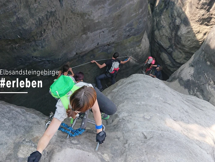 Klettersteigtouren Elbsandsteinguides Personen klettern in einer steilen Felswand im Elbsandsteingebirge, tragen Rucksäcke und nutzen Seile; die Atmosphäre wirkt abenteuerlich.People climb up a steep rock face in the Elbe Sandstone Mountains, carrying rucksacks and using ropes; the atmosphere is adventurous.Lidé lezou po strmé skalní stěně v Labských pískovcích s batohy na zádech a na lanech, atmosféra je dobrodružná.Ludzie wspinają się na stromą ścianę skalną w Górach Piaskowca Łabskiego, niosąc plecaki i używając lin; atmosfera jest pełna przygód.Mensen beklimmen een steile rotswand in het Elbezandsteengebergte, dragen rugzakken en gebruiken touwen; er hangt een avontuurlijke sfeer.Le persone si arrampicano su una ripida parete rocciosa nelle montagne di arenaria dell'Elba, portando zaini e usando corde; l'atmosfera è avventurosa.