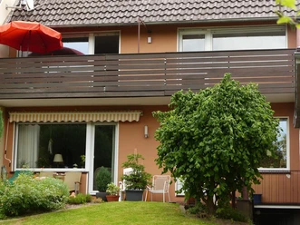 Ein zweistöckiges Wohnhaus mit einem Balkon und einem roten Sonnenschirm. Vor dem Haus steht ein grüner Baum und es gibt einige Pflanzen im Garten.