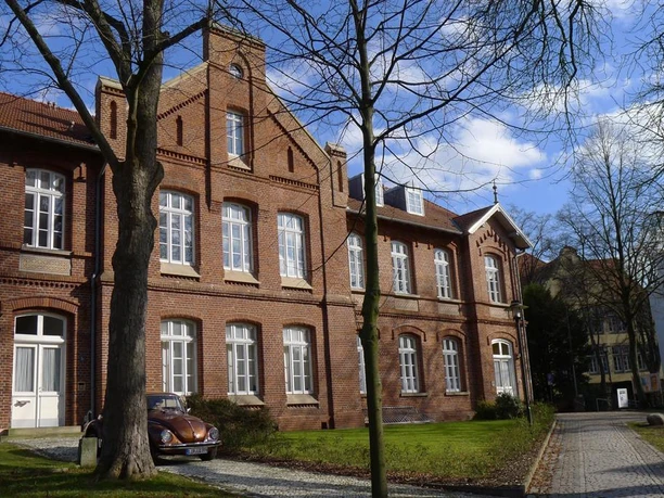 Rotes Backsteingebäude mit weißen Fenstern, Bäumen und gepflasterter Zufahrt