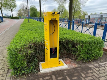 Gelbe ADAC-Radservicestation mit Werkzeugen, umgeben von grünem Gebüsch und nahe einer gepflasterten Straße.