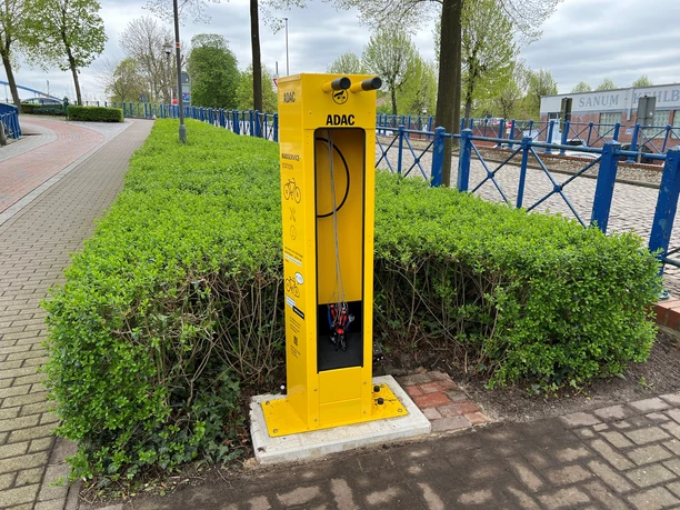 Gelbe ADAC-Radservicestation mit Werkzeugen, umgeben von grünem Gebüsch und nahe einer gepflasterten Straße.