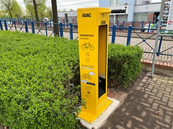 Die gelbe ADAC-Radservicestation steht am Rand eines gepflasterten Gehwegs, umgeben von grünen Hecken.
