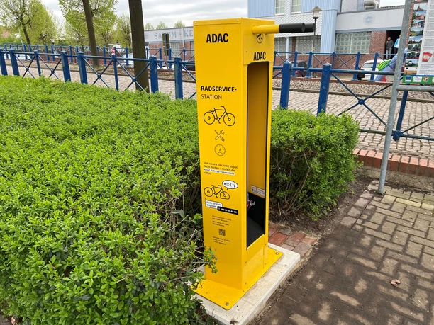 Die gelbe ADAC-Radservicestation steht am Rand eines gepflasterten Gehwegs, umgeben von grünen Hecken.