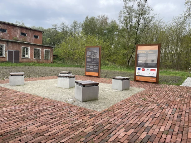 Infotafeln und Sitzblöcke aus Beton auf rotem Pflaster vor altem Backsteingebäude im Grünen