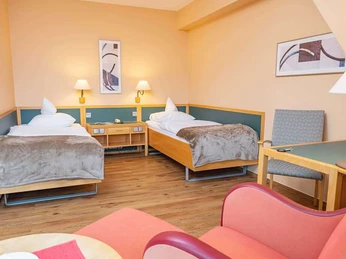 Bad Driburg_Gräfliche Kliniken_Caspar Heinrich Klinik_1024 px.jpg Zwei Einzelbetten in einem hellen Zimmer mit Holzmöbeln und einem Schreibtisch im Vordergrund.