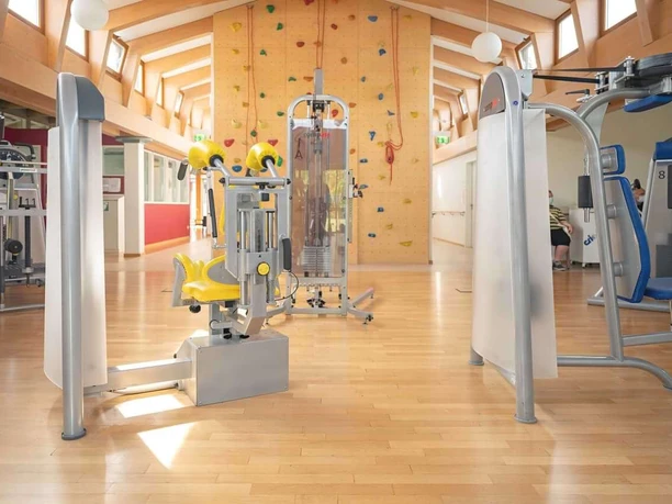 Bad Driburg_Gräfliche Kliniken_Caspar Heinrich Klinik Heller Trainingsraum mit modernen Fitnessgeräten und Kletterwand unter einem Holzgebälk.
