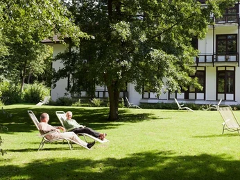 Zwei Personen entspannen auf Liegestühlen im Schatten großer Bäume vor einem Klinikgebäude.