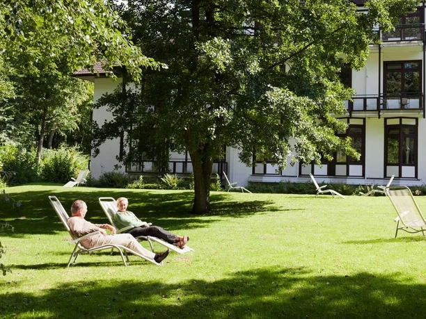 Bad Driburg_Gräfliche Kliniken_Marcus Klinik 3_1280 px.jpg Zwei Personen entspannen auf Liegestühlen im Schatten großer Bäume vor einem Klinikgebäude.
