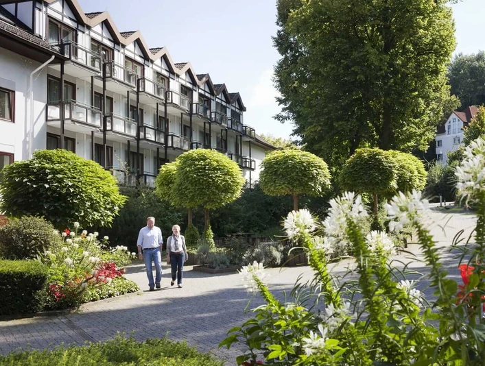 Bad Driburg_Gräfliche Kliniken_Marcus Klinik 2_1280 px.jpg Älteres Paar spaziert durch eine grüne Gartenanlage vor der Marcus Klinik mit blühenden Pflanzen.