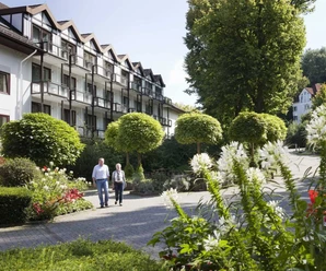 Bad Driburg_Gräfliche Kliniken_Marcus Klinik Älteres Paar spaziert durch eine grüne Gartenanlage vor der Marcus Klinik mit blühenden Pflanzen.