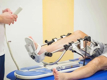 Ein Patient liegt auf einem Therapiegerät zum Kniebeugertraining, betreut von einer Fachperson.