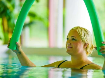 Bad Driburg_Gräfliche Kliniken_Park Klinik 3_1280 px.jpg Frau im Schwimmbecken hält grünen Schwimmstab, umgeben von sanftem Tageslicht und Pflanzen.