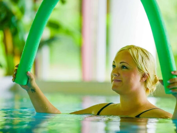 Bad Driburg_Gräfliche Kliniken_Park Klinik 3_1280 px.jpg Frau im Schwimmbecken hält grünen Schwimmstab, umgeben von sanftem Tageslicht und Pflanzen.