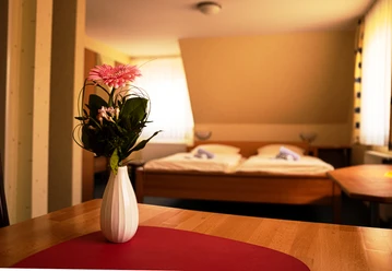 Zweibettzimmer mit Holzinterieur, Blumen in weißer Vase auf Tisch im Vordergrund, Tageslichtfenster.