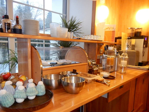 Frühstücksbuffet mit Säften, Obst, Eiern und Gebäck auf Holztheke in einem hellen Raum mit großen Fenstern.