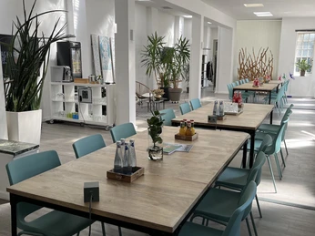 BACKYARD LOFT - Eventlocation Großer, lichtdurchfluteter Raum mit Holz-Sitzgruppen und blauen Stühlen in minimalistischem Design.Large, light-flooded room with wooden seating areas and blue chairs in a minimalist design.