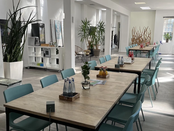 BACKYARD LOFT - Eventlocation Large, light-flooded room with wooden seating areas and blue chairs in a minimalist design.